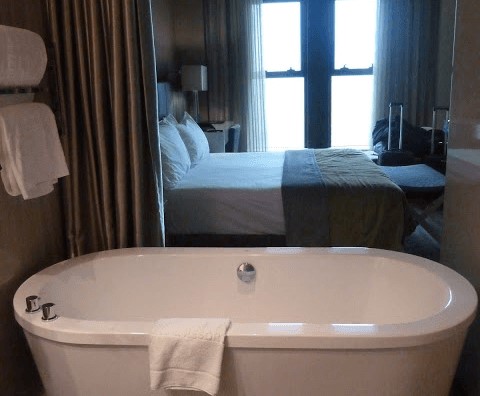 Habitación de hotel en primer plano se ve una bañera blanca y detrás una cama y una ventana con cortinas abiertas