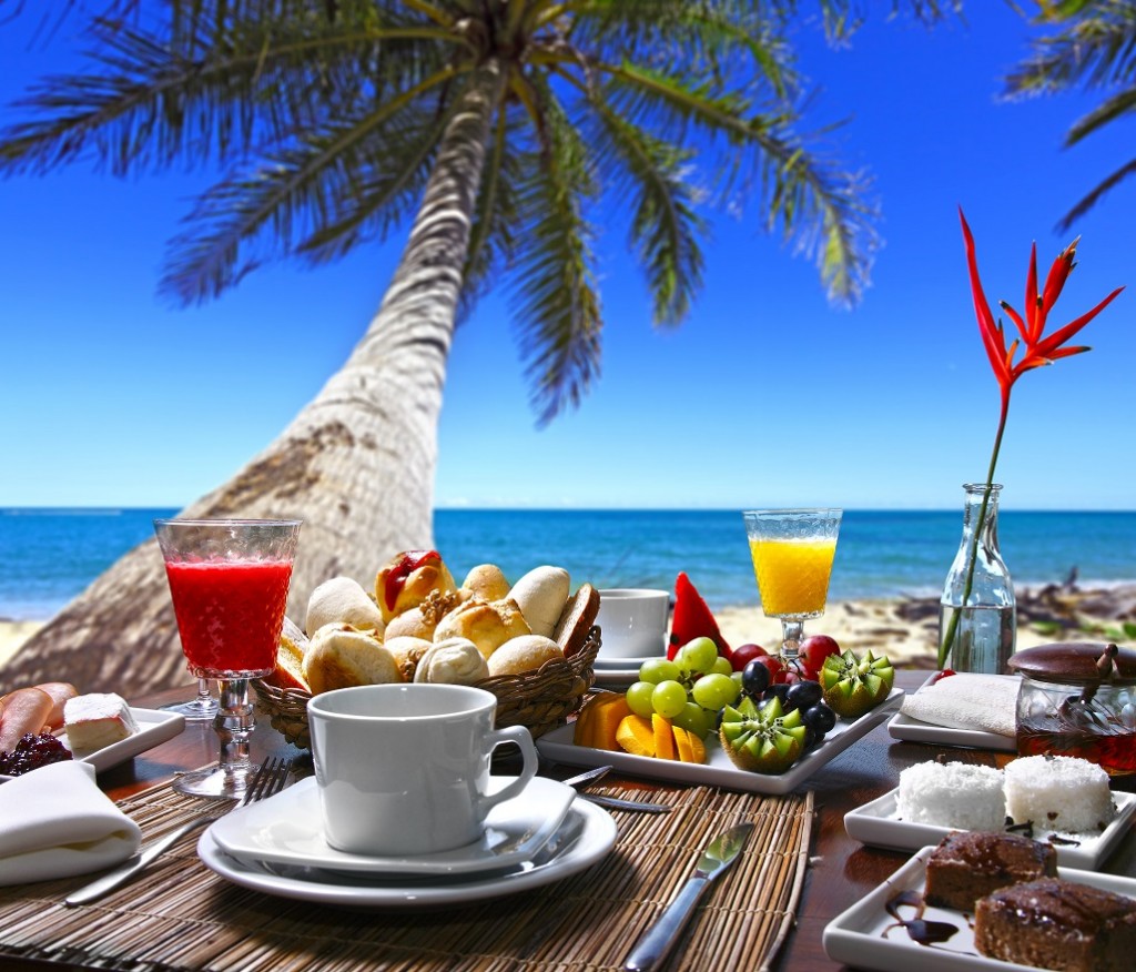 mesa de desayuno con café, pancitos, zumo de fruta, platos de fruta y pasteles, enfrente a la playa con palmeras