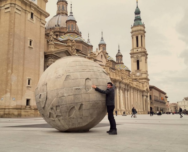 chico abrazando una escultura redonda, similar a un mundo gigante, y detrás la iglesia del Pilar