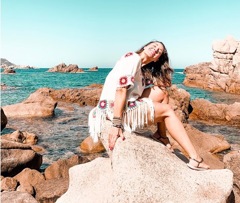 chica posando sentada mirando al sol arriba de una piedra con el mar de fondo